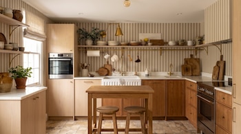 Una cocina con gabinetes de madera clara y oscura, encimeras de mármol, fregadero doble, horno, estufa, mesa y dos taburetes de madera, y estantes con objetos.