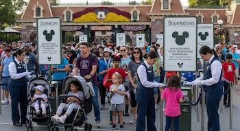 Una fila de visitantes en la entrada de Disneyland, con carteles sobre reconocimiento facial, cámaras y personal del parque asistiendo a familias con niños pequeños.