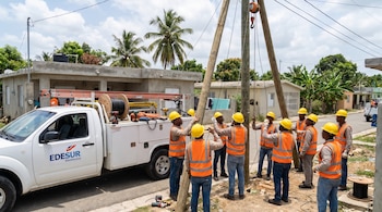 Obreros con casco amarillo y chaleco naranja instalan postes de madera y cableado cerca de un camión blanco con logo Edesur Dominicana en zona urbana con casas.