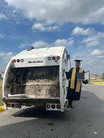 Un hombre con toga, birrete y estola de graduación sostiene su título junto a un camión municipal blanco con la puerta abierta