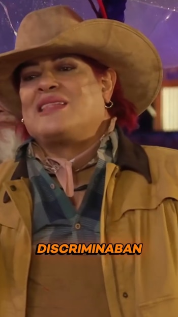 Pimpinela Escarlata viste sombrero vaquero, bufanda y chaqueta beige, con la palabra DISCRIMINABAN escrita abajo