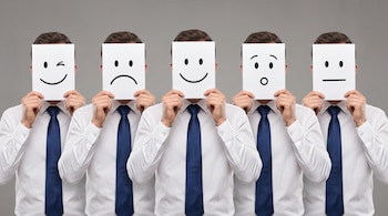 Cinco hombres en camisa blanca y corbata azul sostienen máscaras de papel con emojis de guiño, tristeza, alegría, sorpresa y neutralidad sobre sus caras.