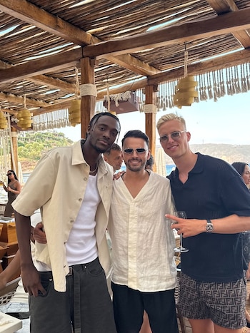 Tres hombres sonriendo a la cámara en un entorno de bar de playa con un techo de madera y el mar de fondo. Uno de ellos sostiene una copa de vino
