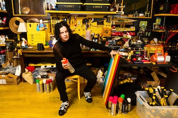 Jack White, vestido de negro, sentado en un taburete amarillo en un taller desordenado, rocía pintura de una lata mientras sostiene listones de madera de colores