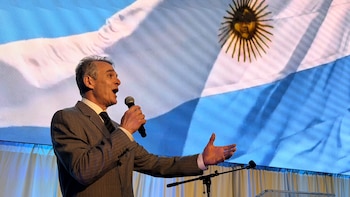 Daniel Bouchet, un hombre de cabello gris con traje a rayas, canta en un micrófono. Detrás, se proyecta la bandera argentina con el Sol de Mayo