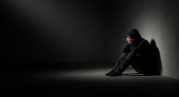 Un adolescente se encuentra agachado en la esquina de un cuarto oscuro, cubriéndose el rostro con las manos, vestido con sudadera y pantalones.