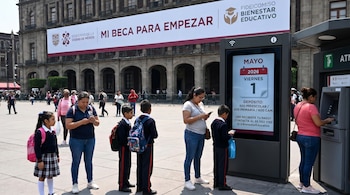 Personas y estudiantes en uniforme escolar frente a una pantalla informativa y un cajero automático en una plaza pública con un gran banner de Mi Beca para Empezar.