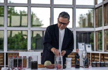 Hombre con gafas y chaqueta negra junto a una mesa con recipientes, cables, batata, melón, pan, botella de vino tinto, copa y medidor eléctrico