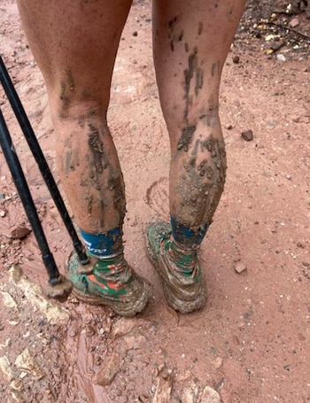 Piernas de una persona cubiertas de barro con zapatillas deportivas, calcetines y bastones de senderismo sobre un terreno embarrado