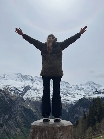Mujer de espaldas con chaqueta verde y pantalones negros de campana, brazos extendidos, de pie sobre un tronco, mirando montañas nevadas y valles verdes bajo un cielo nublado