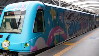 Un tren de metro azul decorado con gráficos de colores, la frase Happy Jiminday y la caricatura de un personaje amarillo