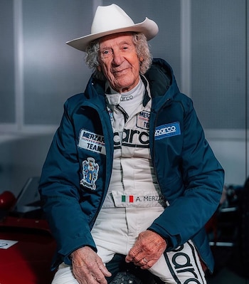 Hombre mayor con cabello rizado gris y sombrero de vaquero blanco. Viste chaqueta azul y traje de carreras blanco con logos Sparco, Merzar Team y bandera italiana