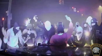 Una multitud de personas se congrega por la noche; varias llevan pasamontañas o capuchas, algunas levantan los brazos o sostienen teléfonos bajo luces brillantes