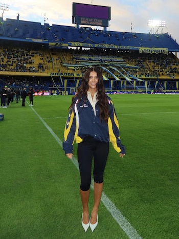Zaira Nara sonríe de pie en el césped de La Bombonera, con chaqueta azul y amarilla, camisa blanca, pantalones negros y tacones blancos. Fondo de gradas azules y amarillas del estadio