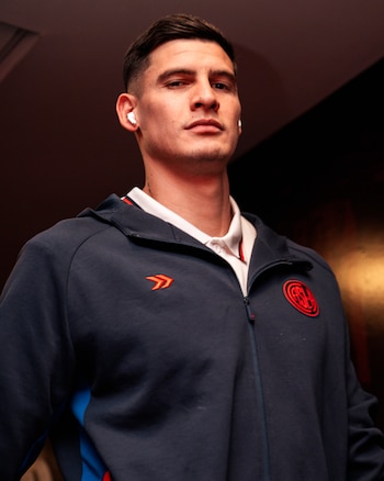 Hombre joven de cabello oscuro y barba incipiente. Viste sudadera azul marino con cremallera y polo blanco, lleva auriculares inalámbricos blancos y dos logos rojos