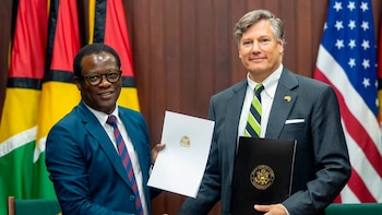 Dos hombres vestidos con traje se dan la mano. Un hombre sostiene un documento blanco; el otro, una carpeta negra. Banderas de Guyana y de Estados Unidos de fondo