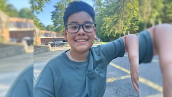 Primer plano de un joven con gafas y camiseta verde sonriendo; árboles y un edificio borroso al fondo