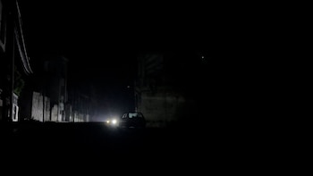 Calle oscura de noche con un automóvil en el centro que ilumina el camino con sus faros. Edificios y postes de luz son apenas visibles