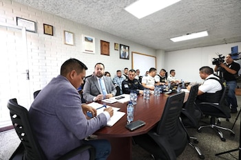 Personas vestidas de traje y policías uniformados sentados en una mesa de conferencia mientras dos personas graban con teléfonos