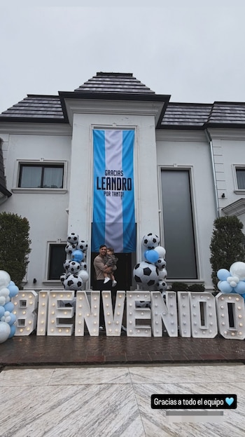 Un hombre sostiene a un niño pequeño frente a una casa. Una bandera argentina cuelga de la fachada y letras luminosas deletrean 'BIENVENIDO'. Hay globos de fútbol y celestes