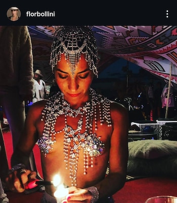 Una mujer, Flor Bollini, usa un tocado y joyas brillantes en cabeza y torso mientras enciende una vela con un mechero, con una tela estampada de fondo