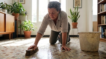 Mujer arrodillada friega un piso de mosaico con un cepillo, mientras una cubeta de plástico sucia está a su lado. La habitación está ordenada e iluminada.