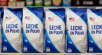 Fila de paquetes blancos y azules de un kilogramo de leche en polvo con una bandera de Estados Unidos en cada uno, sobre un estante de supermercado.