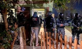 Varios policías con equipo táctico y una persona de civil frente a la entrada de una casa de noche. Hay vegetación, una cerca de madera y un auto
