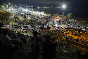 Imagen nocturna desde una elevación con cientos de personas, vehículos, carpas y farolas en una zona costera, con luces de ciudad al fondo