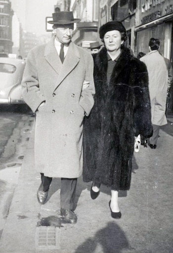 Imagen en blanco y negro de Catherine Dior y Hervé de Charbonneries caminando del brazo. Él usa sombrero y abrigo claro; ella, abrigo de piel oscuro y gorra