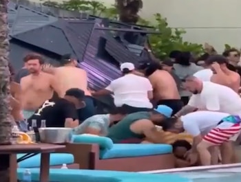 Várias pessoas perto de uma piscina, algumas se abaixando para ajudar outras. Uma estrutura metálica danificada pode ser vista ao fundo. Existem espreguiçadeiras e mesas