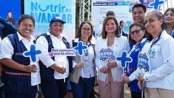Un grupo de seis personas, en su mayoría mujeres, sonríen mientras sostienen carteles con cruces y un autobús azul frente a un letrero con "Nutrir es Avanzar"