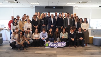 Gran grupo de hombres y mujeres de diversas edades posando para una foto grupal en una oficina moderna. Algunos sostienen un cartel con el hashtag #SoyBP