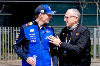 Stefano Domenicali, con gafas y chaqueta negra, conversa con Max Verstappen, quien lleva una gorra y chaqueta azul de Red Bull Racing y sostiene una lata