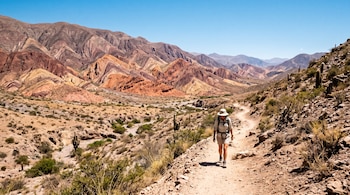 Una persona con sombrero y mochila camina por un sendero pedregoso en una zona árida con cerros de distintos colores de fondo.