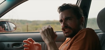 Un hombre con barba y camiseta naranja gesticula con las manos levantadas dentro de un automóvil