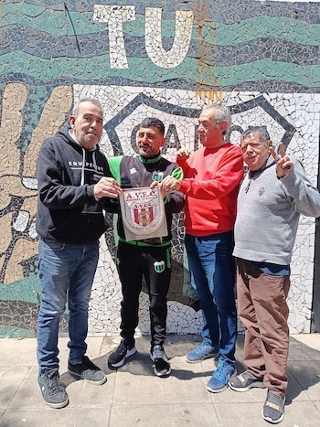 Cuatro hombres adultos sostienen un banderín frente a una pared decorada con mosaicos y letras