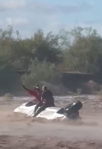 Un hombre se encuentra sentado sobre la base de un vehículo blanco volcado en un río de agua turbia rodeado de vegetación