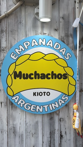 Un cartel ovalado azul y amarillo de "Empanadas Argentinas Muchachos KIOTO" está montado sobre una pared de tablas de madera envejecida