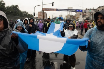 Personas con impermeables y capuchas caminan por la calle mojada y sostienen dos banderas celestes y blancas de gran tamaño