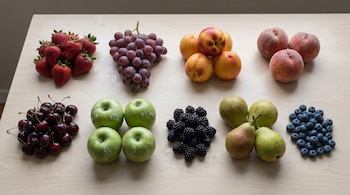 Frutas frescas variadas, como fresas, uvas, nectarinas, melocotones, cerezas, manzanas, moras, peras y arándanos, organizadas en grupos sobre una mesa de madera.