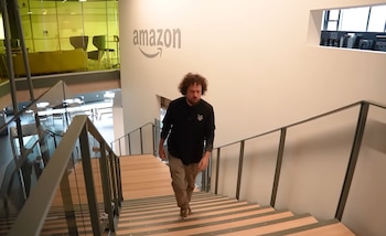 Hombre con cabello rizado baja por escaleras de madera con barandales de metal y cristal. Pared blanca con logo de Amazon. Espacio de oficina con sofás y mesas