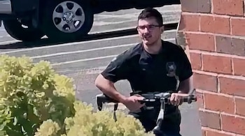 Hombre con gafas y camisa oscura sujetando un fusil, un muro de ladrillo, arbustos verdes en primer plano y la rueda de un vehículo.