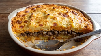 Bandeja de cerámica con un pastel de papas y batata con capas de puré y carne, superficie gratinada y una cuchara metálica. Fondo de madera.