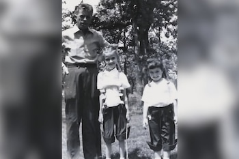 Fotografía en blanco y negro de un hombre y dos niñas posando al aire libre; la niña central lleva un soporte ortopédico y un bastón, con árboles de fondo