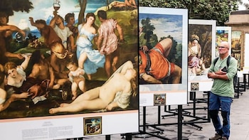 Hombre con gafas y camiseta verde de pie frente a grandes reproducciones de pinturas clásicas al aire libre. Las obras muestran escenas con múltiples figuras humanas