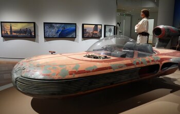 Un vehículo espacial descapotable y una figura con túnica beige están en una sala de museo frente a cuadros colgados en la pared