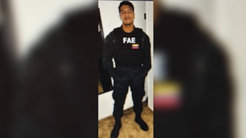 Un hombre de pie, de tez morena, viste un chaleco táctico negro con las siglas FAE y una bandera venezolana, pantalones oscuros y zapatos negros, mirando al frente