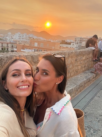 Dos mujeres posan para una selfie; una sonríe y la otra besa su mejilla. De fondo se observa un muro de piedra, un pueblo y un atardecer con montañas
