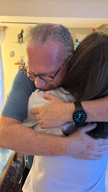 Un hombre con gafas abraza a una mujer de cabello oscuro que está de espaldas en el interior de una vivienda